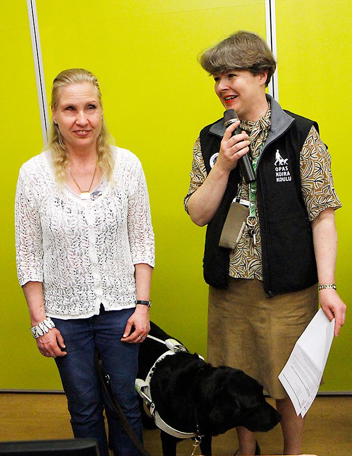 Marianne Tenhami seisoo Iiriksen limenvihreän seinän edessä mustan koiransa Pekon kanssa. Hänen vieressään seisoo Johanna Reiman, joka katsoo Mariannea, puhuu mikrofoniin ja pitää vasemmassa kädessään jotain papereita. Tenhamilla on valkoinen neulepaita ja Reimanilla musta Opaskoirakoulun merkkiä kantava liivi.