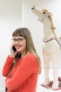 Mari Tammisaari pitää puhelinta korvallaan ja hymyilee kameralle. Taustalla on iso valkoinen hevospatsas, jossa on kukkakoristeita.