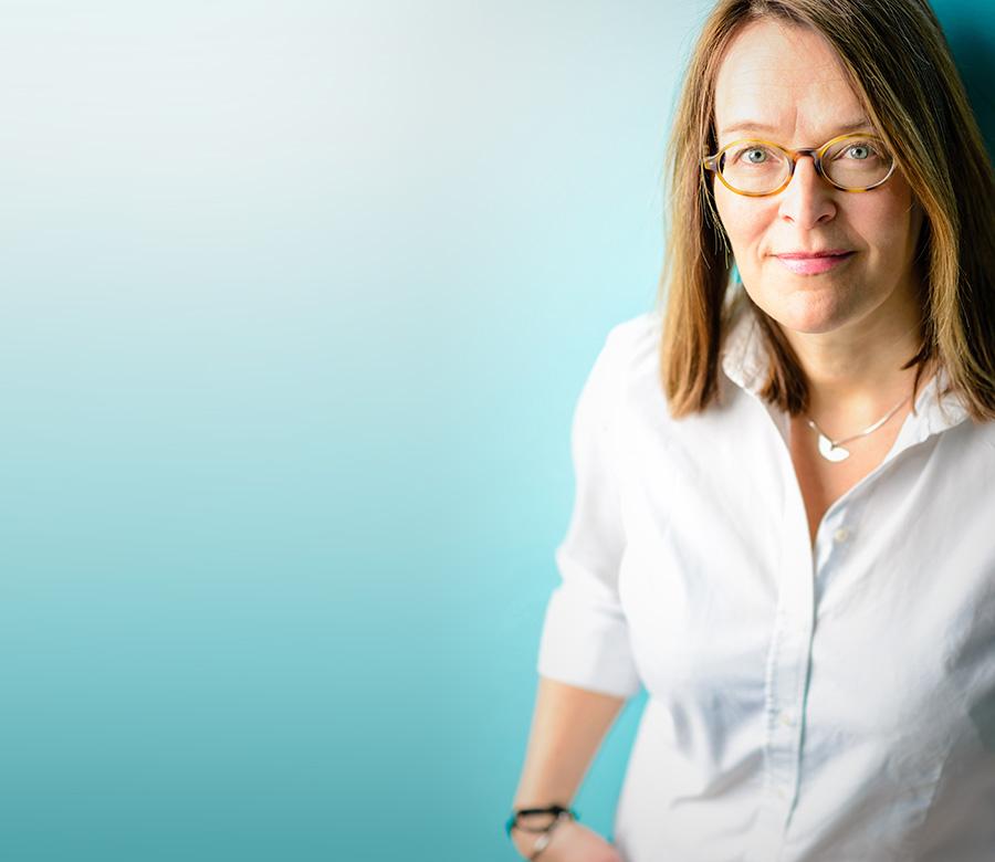 Päätoimittaja Marika Mäkinen valkoisessa paidassa turkoosin seinän edessä. Kuvaan tulee pehmeä ja raikas luonnonvalo.