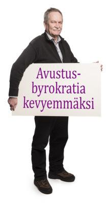 Kari Vähänen pitelee kainalossaan isoa kylttiä, jossa lukee: "Avustusbyrokratia kevyemmäksi".