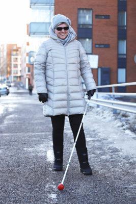 Kristiina Suominen kävelee hymyillen jalkakäytävällä valkoinen keppi kädessään.