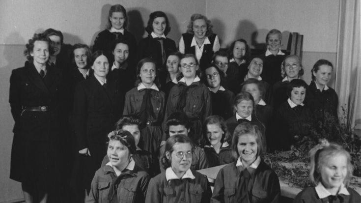 Mustavalkoisessa kuvassa parikymmentä iloista tyttöä partiopuvuissa sekä muutama aikuinen nainen.