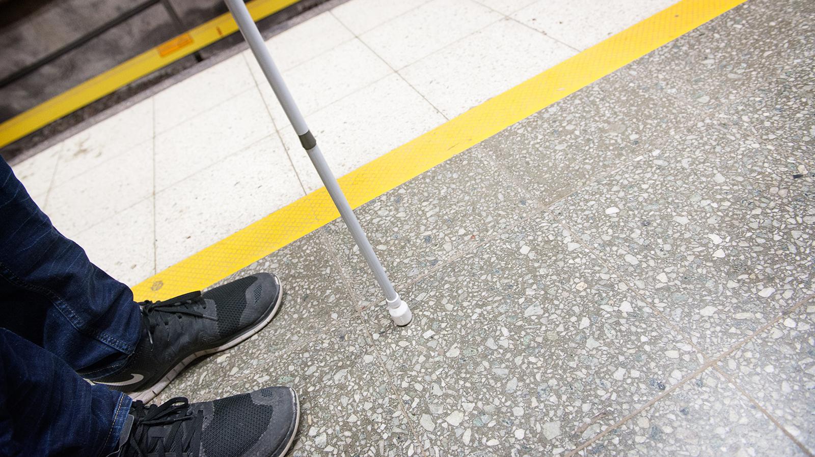 Kuvassa näkyy valkoinen keppi ja sen käyttäjän musta lenkkitossu ja farkun lahje. Maassa näkyy metroaseman lattian keltaista kontrastiraitaa.