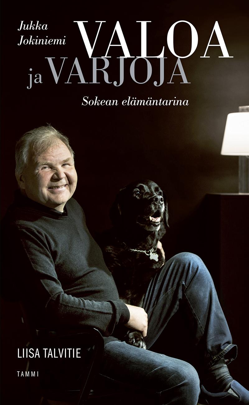 Valoa ja varjoja -kirjan kansikuva. Jukka Jokiniemi hymyilee lampun valossa musta labradorinnoutaja vierellään.
