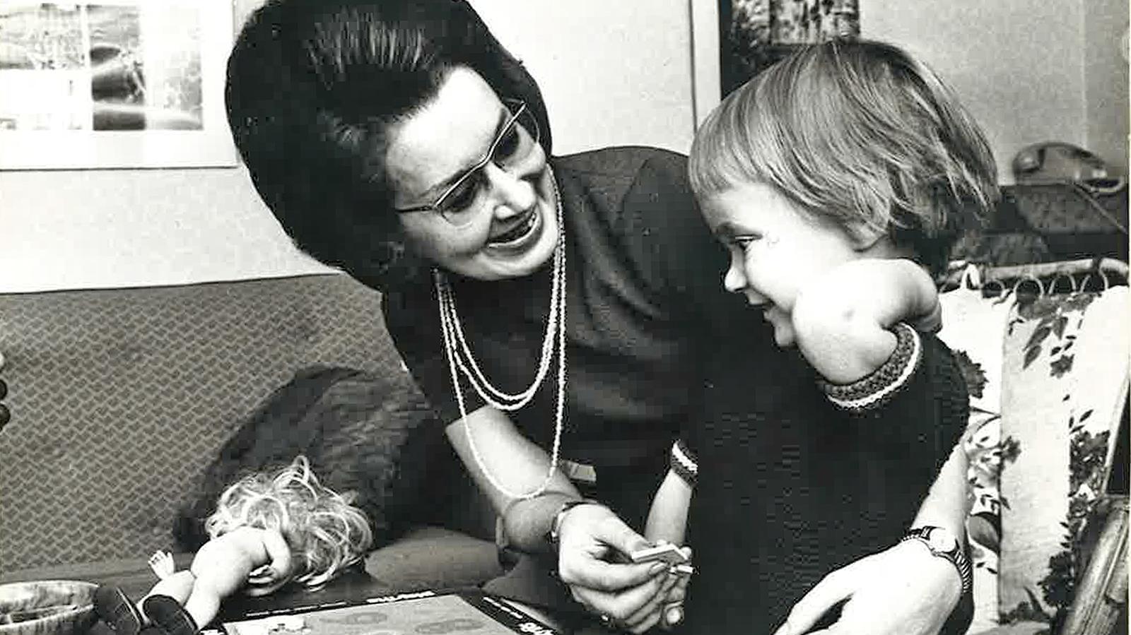 Mustavalkoisessa kuvassa Leena katsoo hymyillen pientä tyttöä, toinen käsi tytön ympärillä ja toisessa kädessä kohonumeropelin palanen.