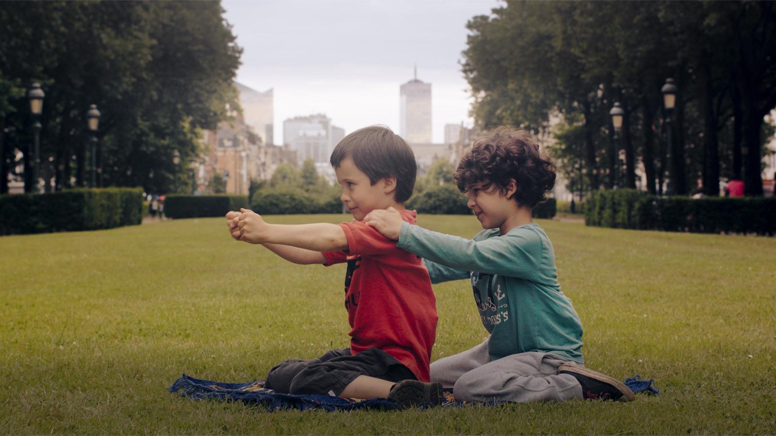 Kaksi poikaa istuu puistossa peräkkäin nurmikolla polvillaan. Taaimmaisen kädet ovat etummaisen hartioilla. Etummaisen kädet ovat suorana edessä yhdessä.