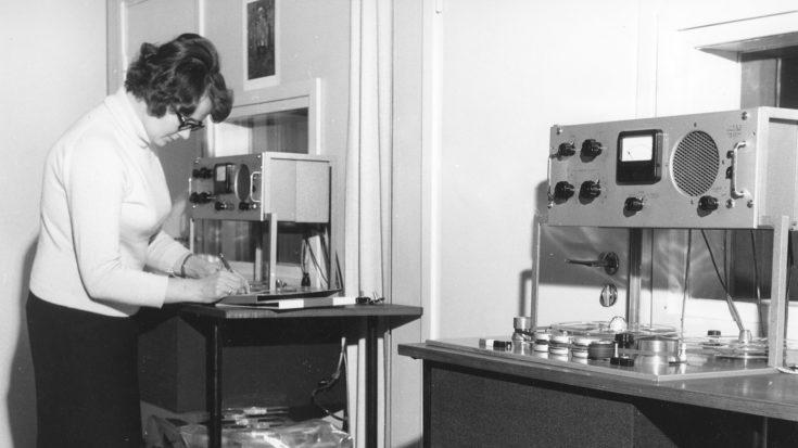 Mustavalkoisessa 60-luvun kuvassa Mirja Kallio kirjoittamassa jotain muistiin äänityslaitteen edessä. Hänellä on valkoinen pitkähihainen pusero ja musta hame. Hiukset ovat tummat, lyhyet ja kihartuvat, ja hänellä on mustasankaiset silmälasit.