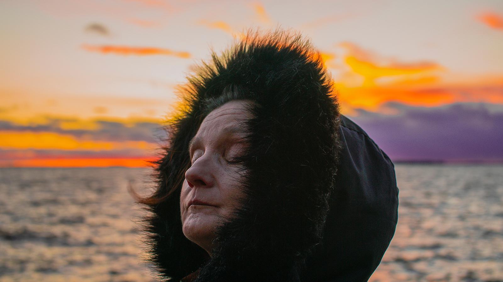 Anneli Iltanen silmät kiinni musta karvareunuksinen huppu päässä, auringonlasku taustalla.
