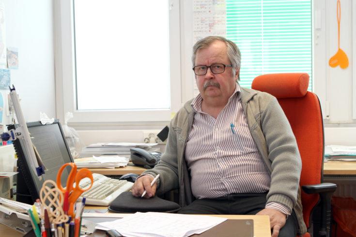 Silmälasipäinen, harmaahiuksinen mies istuu työpöytänsä ääressä, päällään raidallinen kauluspaita ja neuletakki.