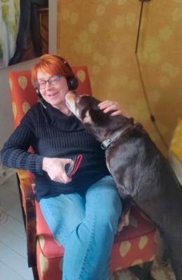 Liisa Riste istuu tuolissa kuulokkeet päässään ja kännykkä kädessään. Musta koira kurottauttuu nuolemaan Risten kasvoja.