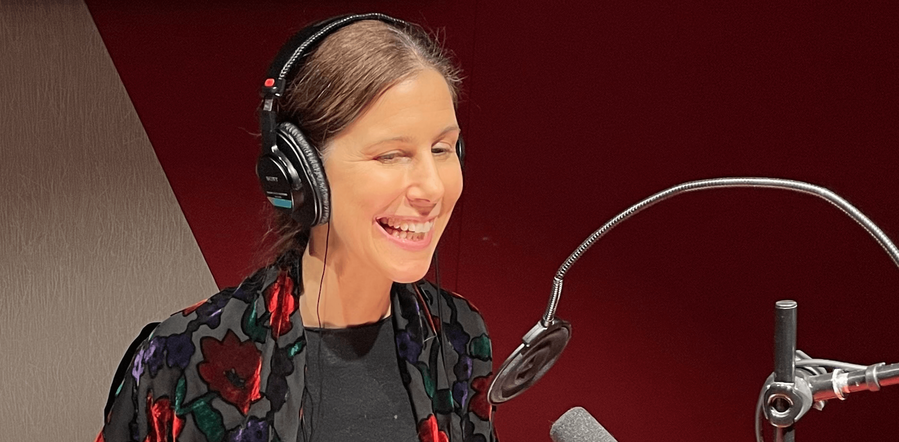 Ruskeahiuksinen, hymyilevä nainen kuulokkeet korvilla ison studiomikrofonin edessä.