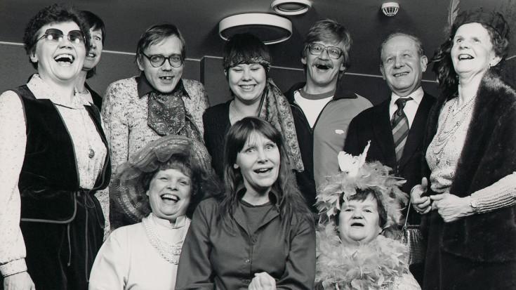 Mustavalkoisessa ryhmäkuvassa kymmenen ihmistä, jotka nauravat.