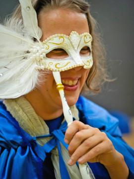 Nainen hymyilee ja pitää kasvoillaan valkoista koristeellista silmät peittävää naamiota, jossa on kultaisia koukeroita, helmiä ja sulkia. Hänellä on sininen viitta harteillaan.