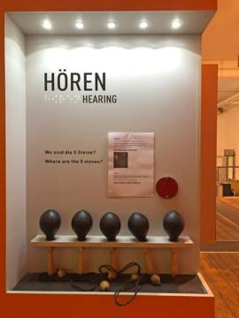 Kuuntelupiste, jossa viisi marakassia ja seinässä teksti "Hören" näkevien kirjoituksella ja pisteillä sekä näkevien kirjoituksella "hearing".