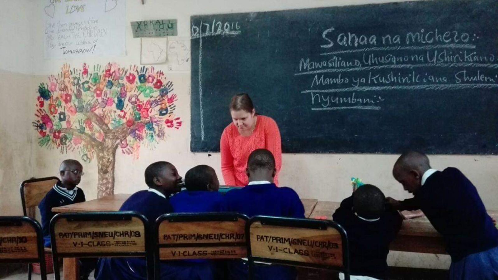 Nainen seisoo liitutaulun edessä. Liitutaululla tekstiä. Edessä kuusi koululaista. Seinällä kuva puusta, jonka "lehtinä" on erivärisiä kädenjälkiä.