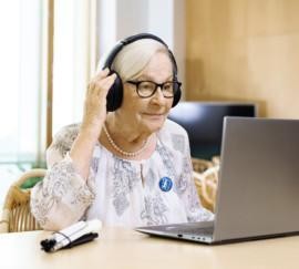 Harmaahiuksinen nainen, jolla on tummasankaiset silmälasit. Päässä kuulokkeet. Edessä pöydällä kannettava tietokone ja kasaan taiteltu valkoinen keppi.