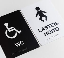 Kaksi kylttiä. Mustassa lukee WC ja siinä on pyörätuolisymboli, valkoisessa lukee lastenhoito ja siinä on yksinkertainen lapsihahmo. Kylttien alareunassa pistekirjoitusta.
