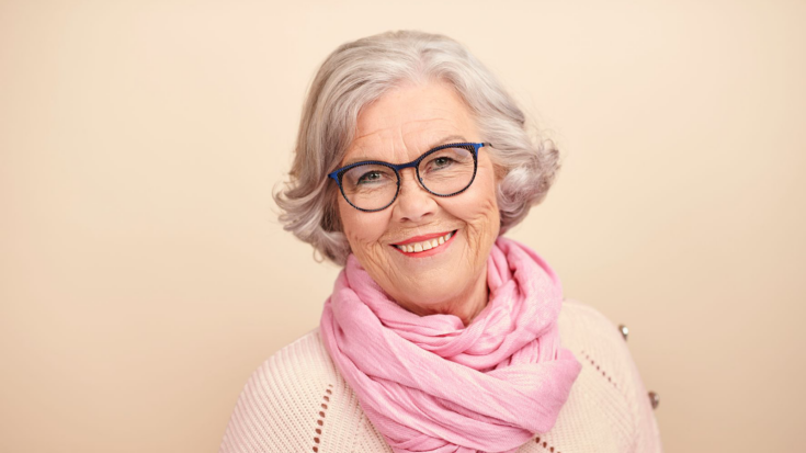 Nainen, jolla on harmaat kiharat poskipäihin ulottuvat hiukset. Tummasankaiset silmälasit. Hymyilee. Kermanvaalea neulepusero ja vaaleanpunainen kaulahuivi.