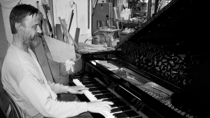 Mustavalkoinen kuva. Parrakas mies soittaa pianoa.
